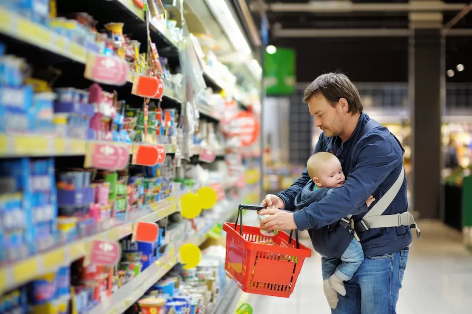 Man with baby in sling holds red grocery basket while looking at refrigerated store section.